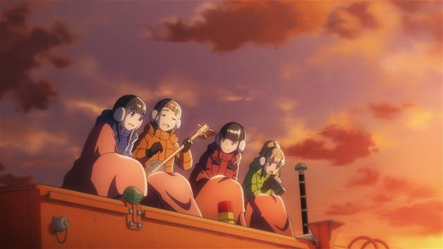 Our four girls sing in sleeping bags on top of a Sno-Cat with earmuffs and coats on as Mari plays a tune on a banjo, sunset lighting the scene a warm orange from the right side of the frame.