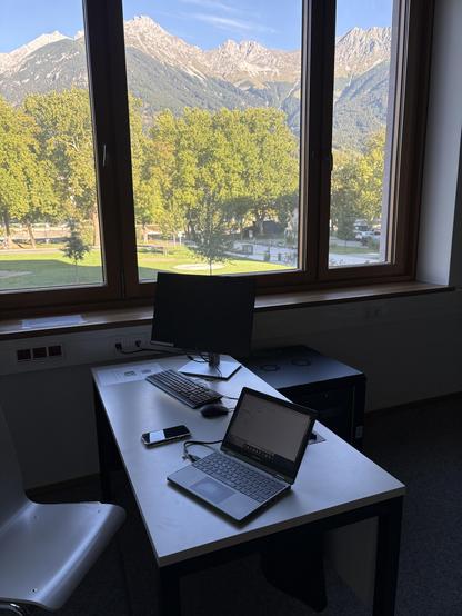 Schreibtisch mit Laptop, dahinter Fenster mit Alpenblick